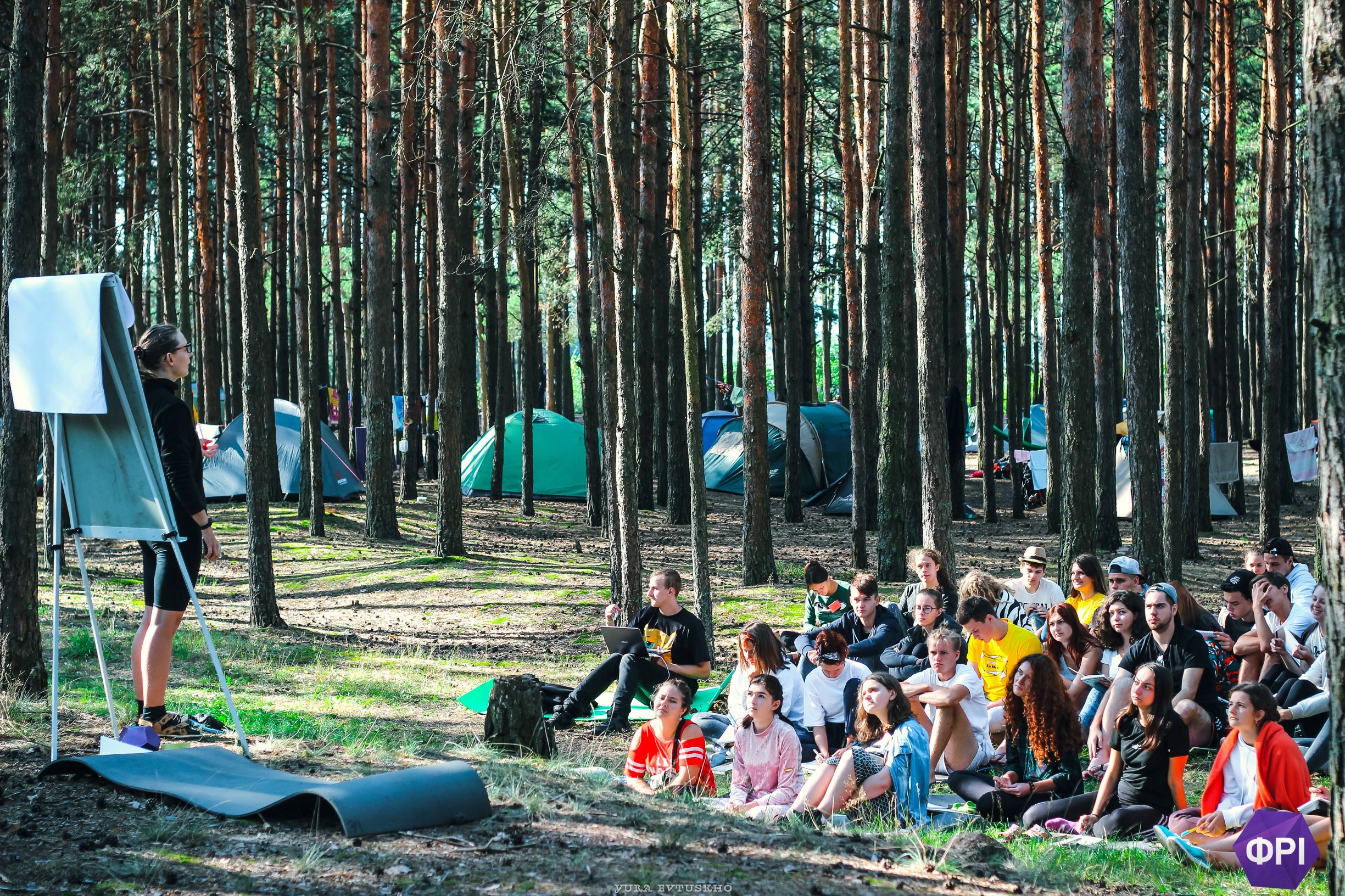 Roman standing next to a tree listening to a lecture. Trees and a lake in the background. Other people in the foreground.
