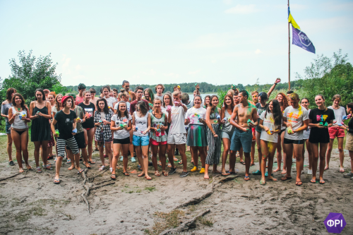 A gif, where approximately thirty people mostly in white shirts throw red, green, blue and yellow holi dyes over themselves and others and get sprayed with color.