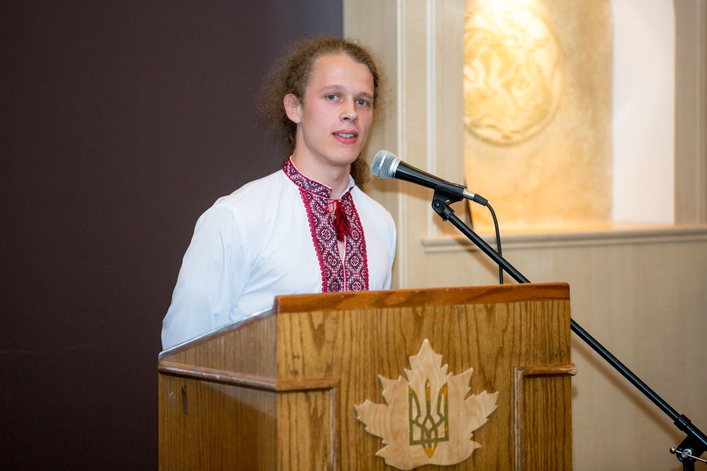 Roman giving a speech at a podium, decorated with the Ukrainian Coat of Arms wearing an embroidered shirt.