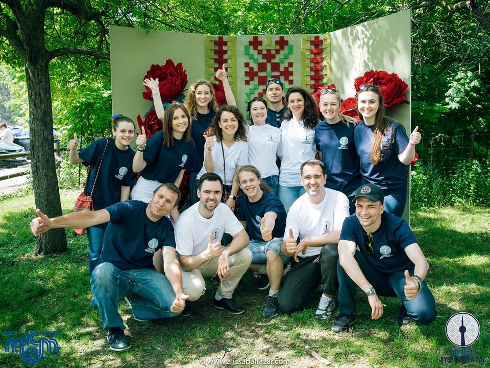 A group of volunteers wearing matching shirts in front of a giant embroidered photo booth.